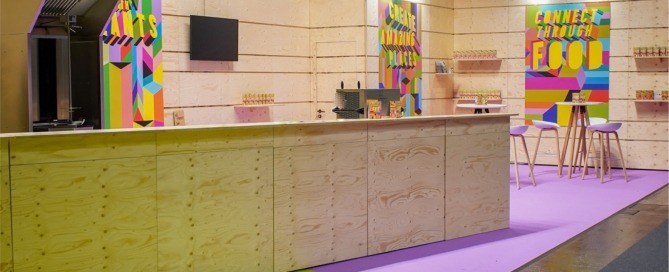 Gastrofläche für die Expo Real 2023, Hochtische und Stühle, Wand mit großen farbintensiven Plakaten und Barbereich
